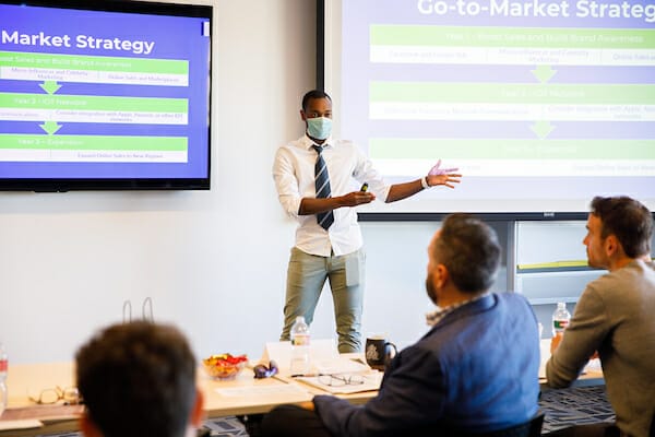 A man wearing a face covering stands and gestures while communicating his idea to a panel of judges and audience.
