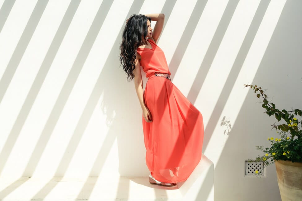 A woman with long, dark hair, wearing an orange dress, leaning against a white wall with stripes of shadows adding interest, and a large potted plant off to the side.