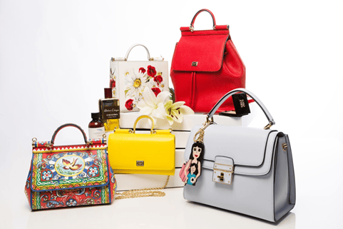 a red and blue folk-print handbag, a yellow handbag, a white, floral print handbag, a red handbag and a gray handbag decorated with a charm of a woman with a child, arranged on flat, white boxes at various heights and accented with lilies and two perfume bottles.