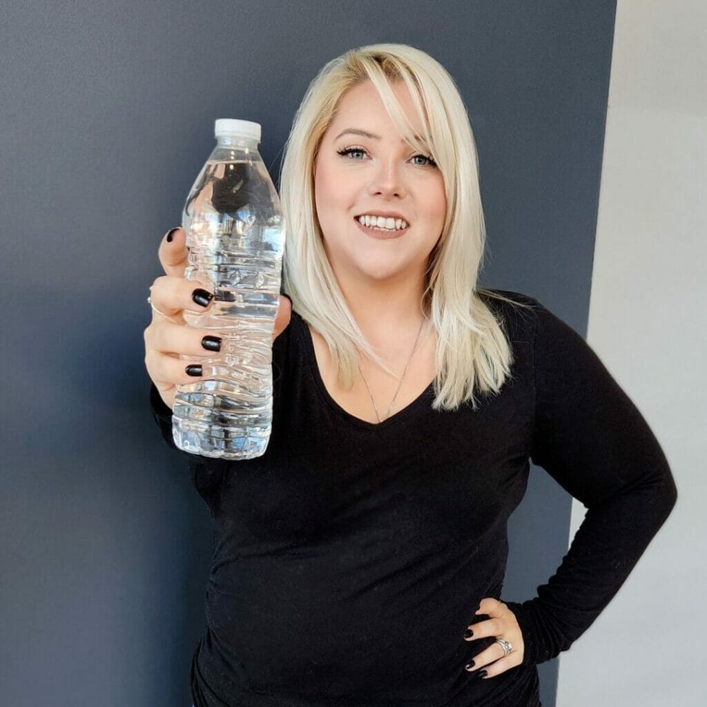 Woman with blonde hair wearing a black top, smiling and holding a water bottle towards the camera.
