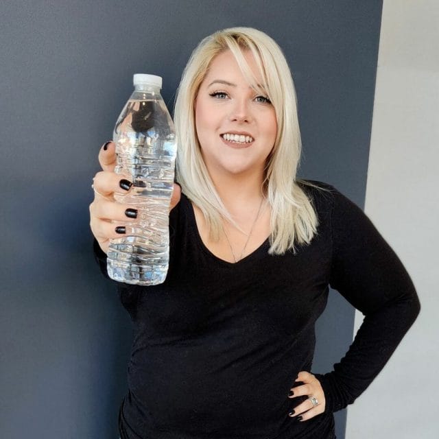 Woman with blonde hair wearing a black top, smiling and holding a water bottle towards the camera.