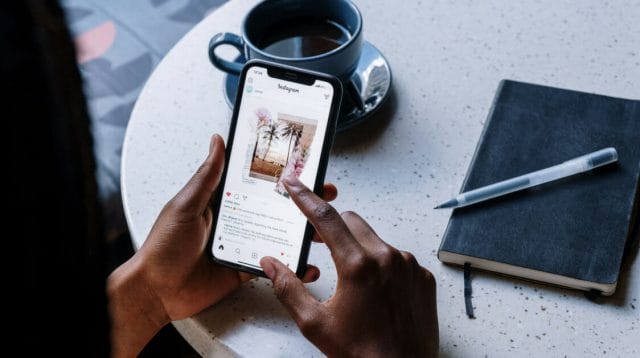 Hands scrolling on a mobile phone with a coffee mug and notebook in the background