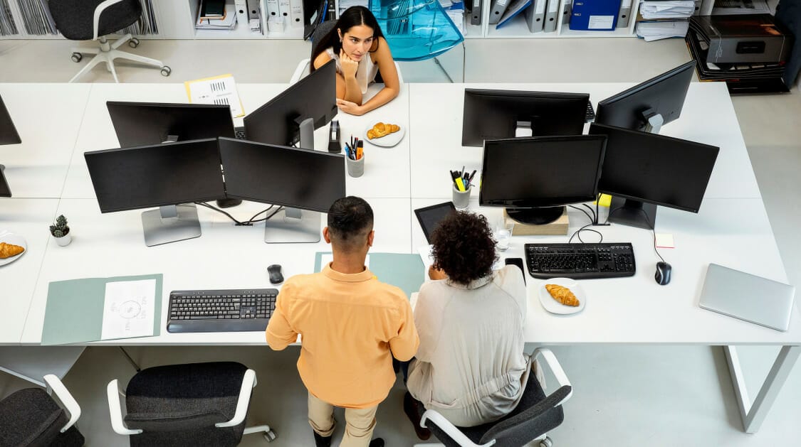 White desks with computer monitors and 3 people working in an office