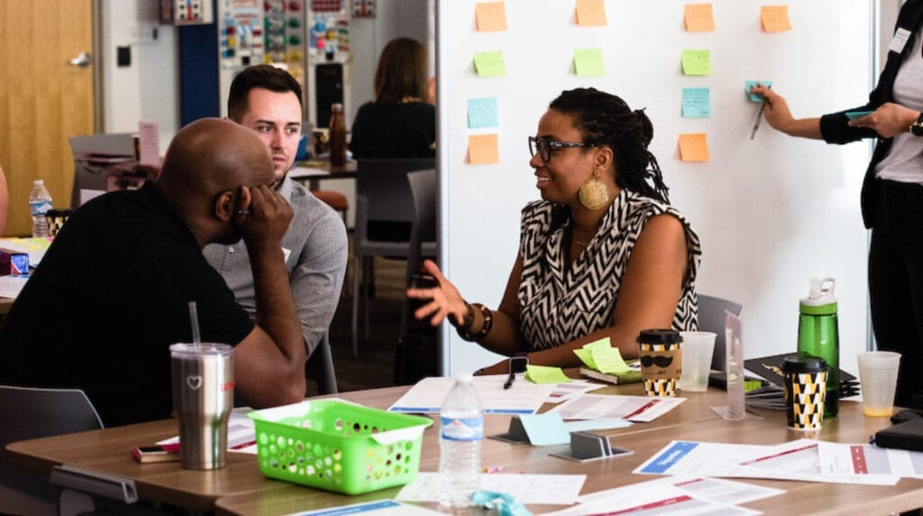 Three people sitting at a table talking to one another with a white board in the background