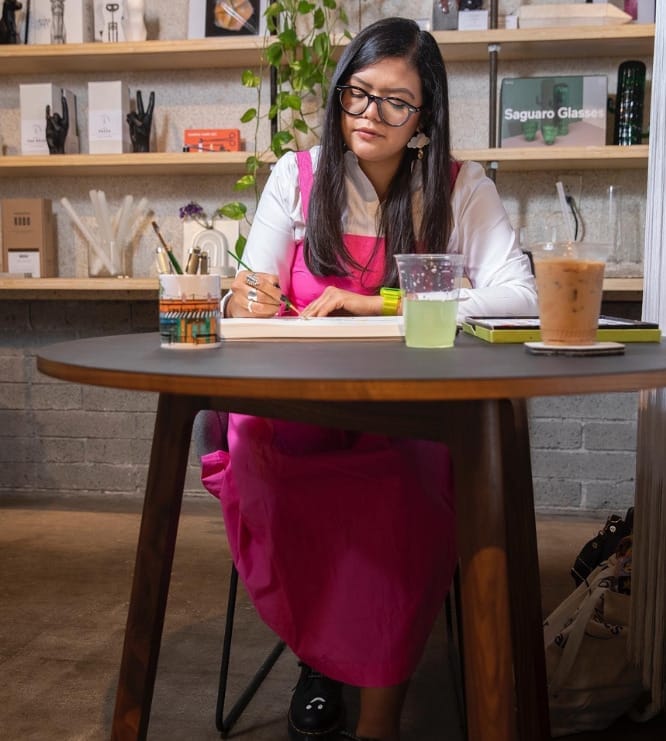 Person sitting and painting at a round table with two cups of liquids and shelves in the background
