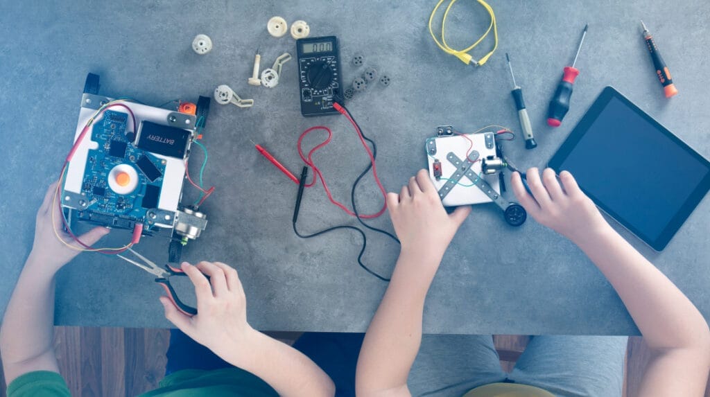 A view looking down at a table with mechanical parts and tools across the table and two children working with the parts and tools.