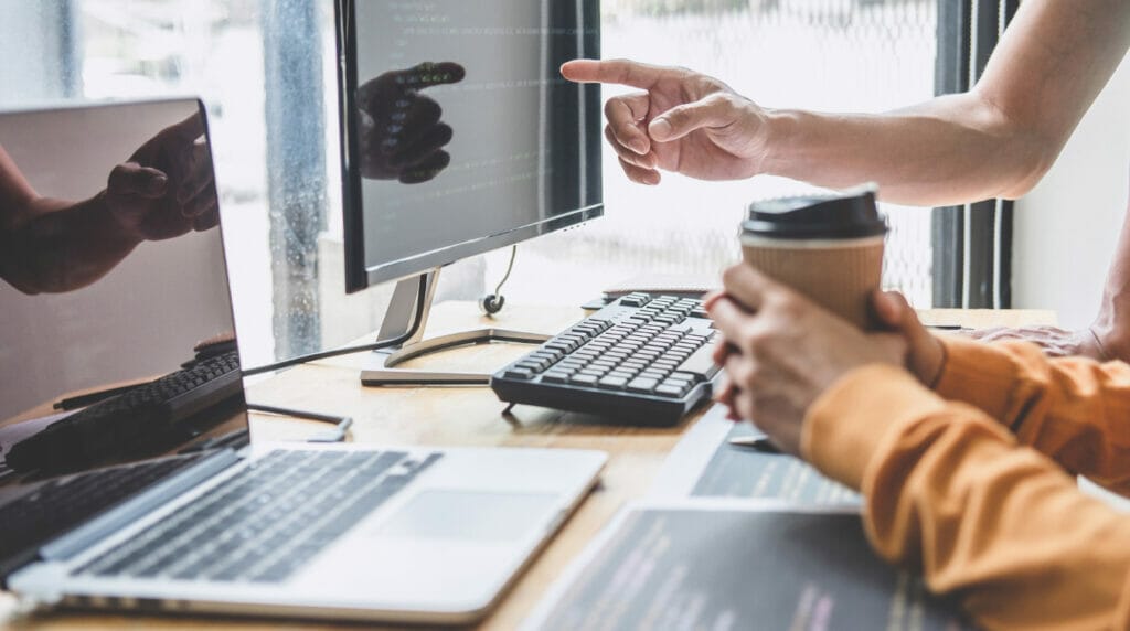 A hand pointing at a computer screen with a laptop on the table and two other hand holding a coffee cup