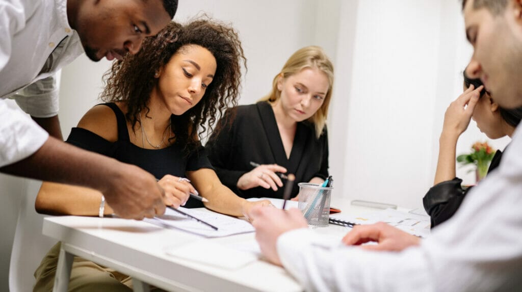  Five professional people sitting at a white table looking at a paper reviewing the content on the paper together