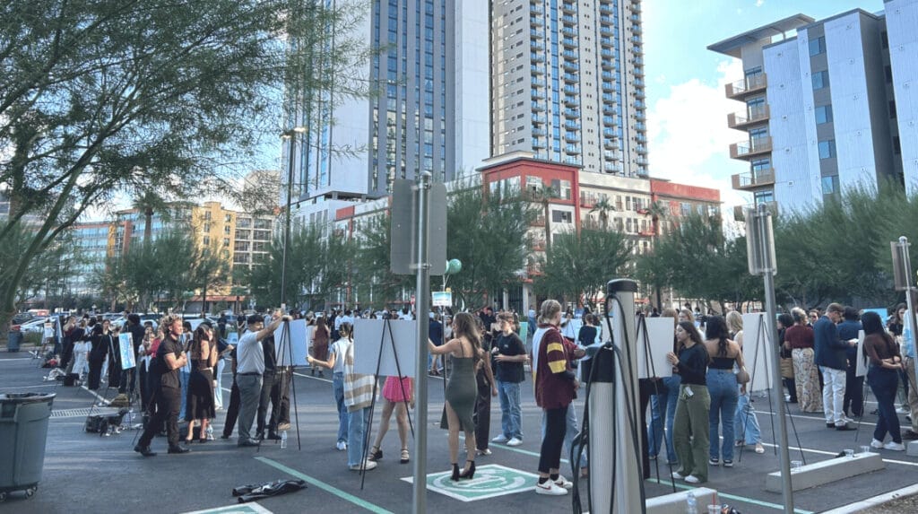 Many people standing outdoors in a parking lot next to presentations on easels with tall buildings in the background