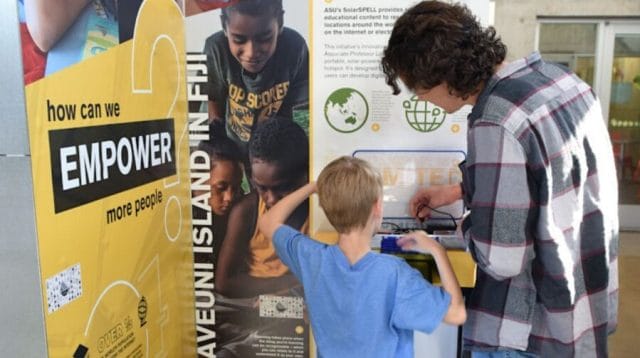 An adult helping a child use manual technology alongside a sign that reads ‘how can we EMPOWER more people?’
