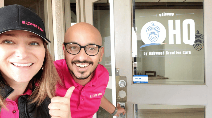 Two people smiling in front of a glass office door, one is wearing a hat and one is wearing glasses