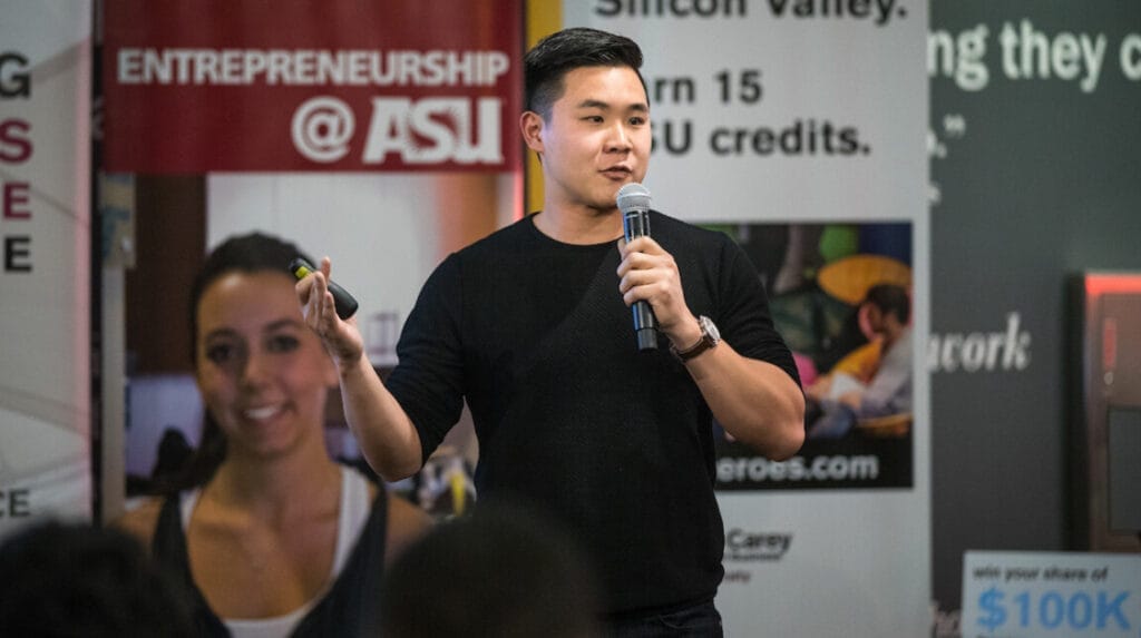 Person in a cark shirt speaking into a microphone with ASU signs in the background