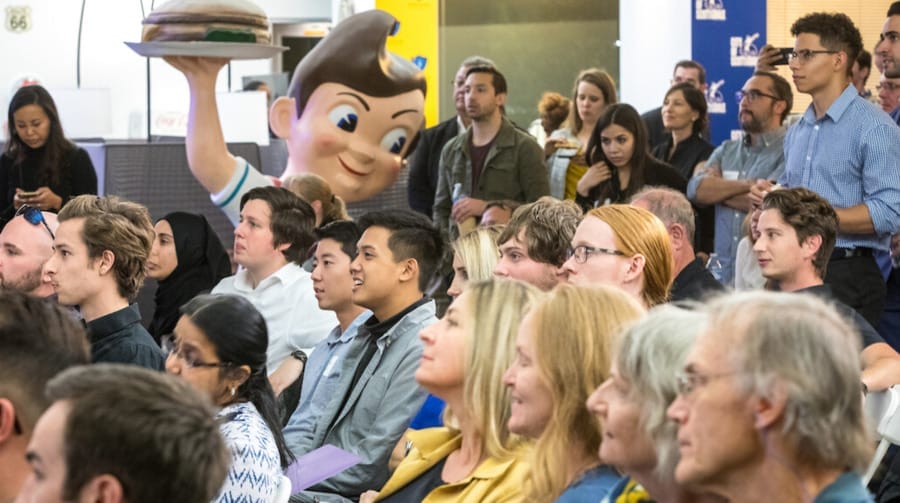 A crowd of people sitting at an event indoors with a vintage statue in the background