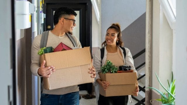 Two students smiling and carrying boxes at their new apartment