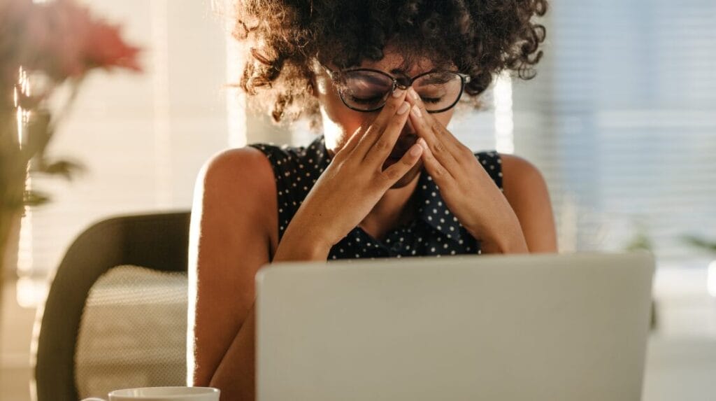 A person wearing glasses, sitting at a desk with their laptop in front of them rubbing their eyes to relieve stress