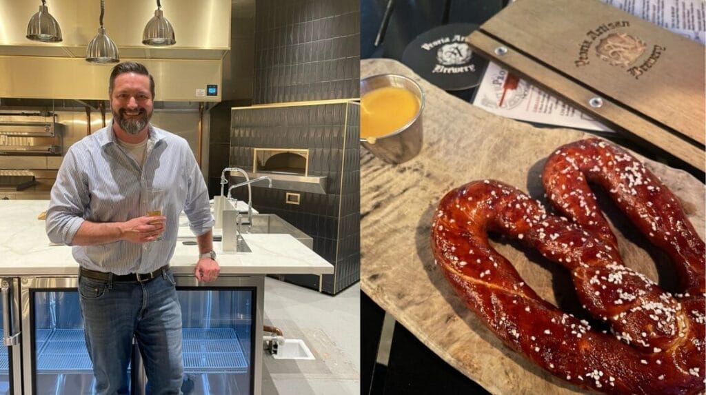 Alt text: A person standing in an upscale restaurant kitchen holding a drink and a large pretzel with cheddar cheese on a table.