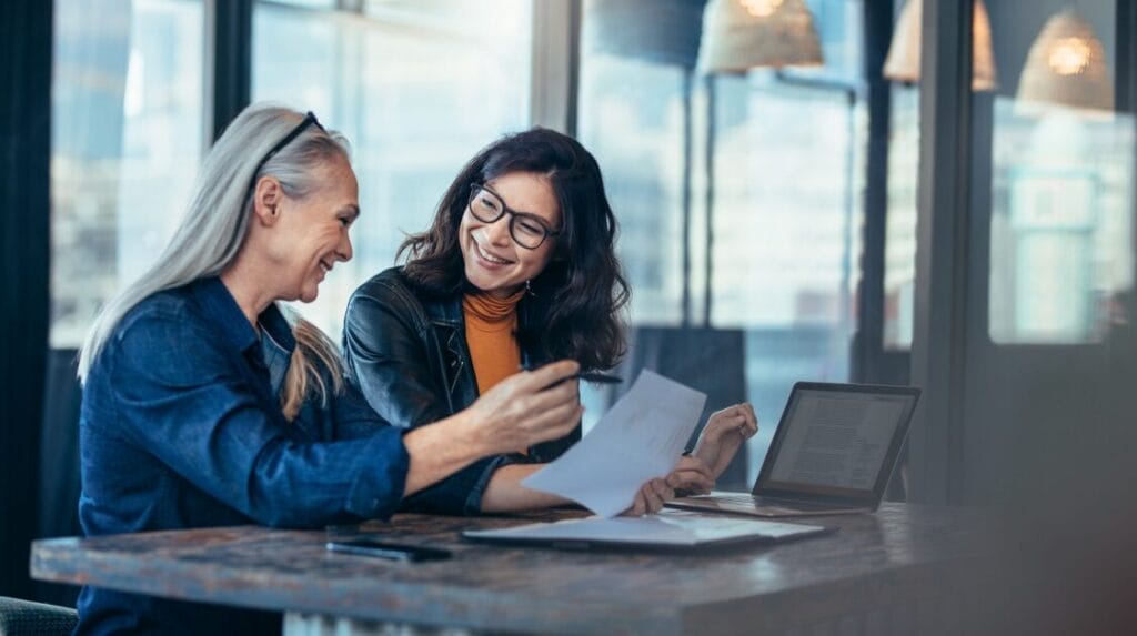 Two people sitting at a table smiling and looking over paperwork together in front of large windows