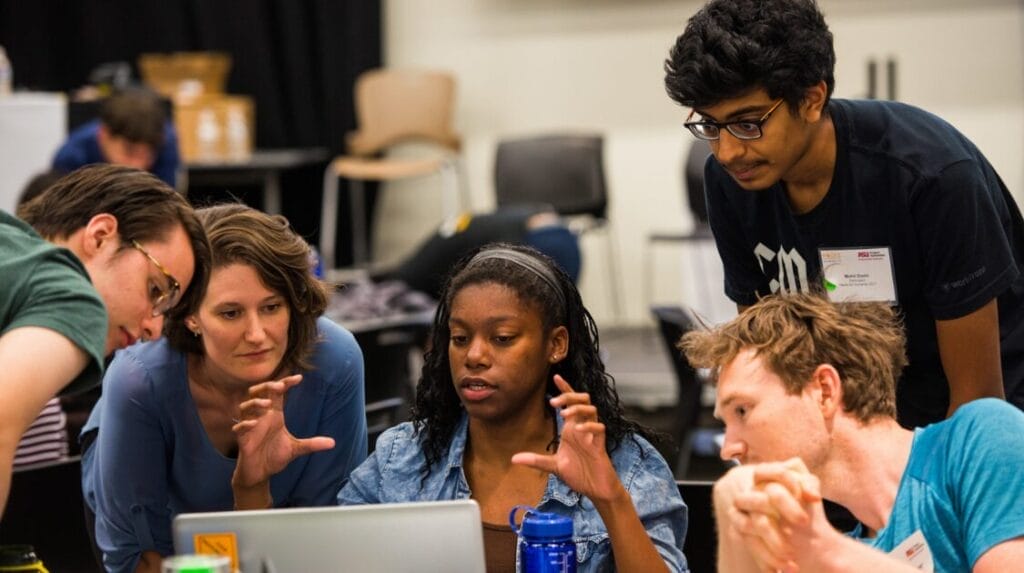 Five people gathered and  looking at a laptop screen as one person shares insights and ideas