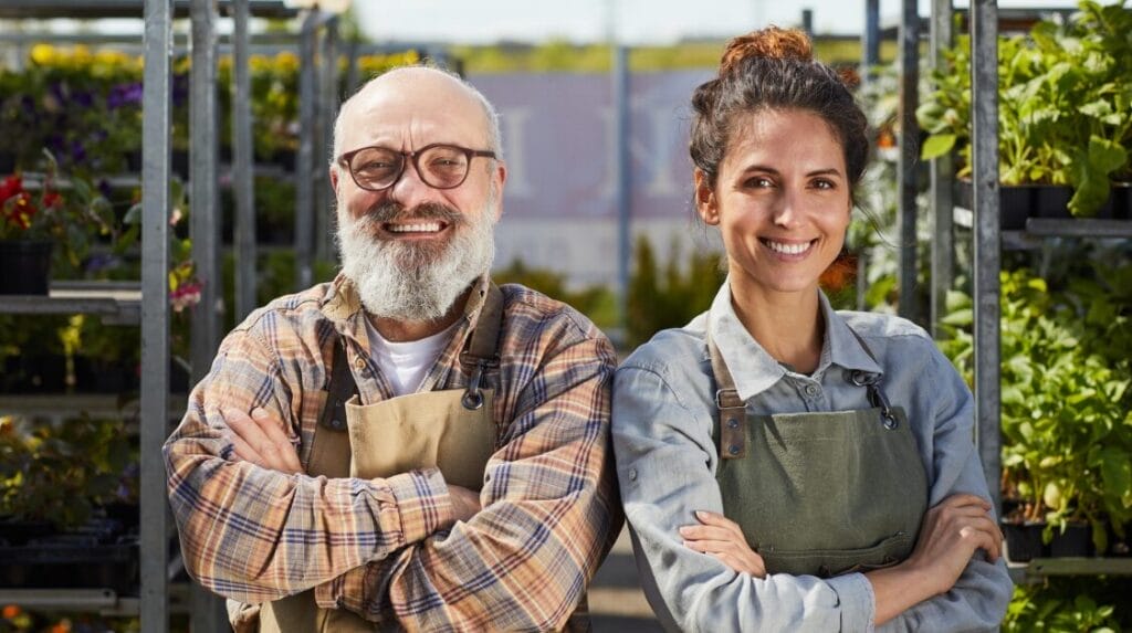 Two people wearing aprons and smiling in front of their gardening business racks