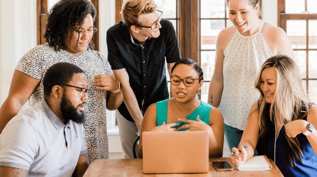 Six people looking at a laptop with bright windows in the background.