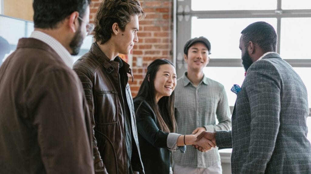 Two people shaking hands with three people standing around them with a brick wall in the background.
