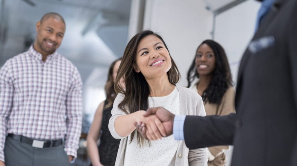 A CEO shakes hands with a business colleague with smiling coworkers in the background