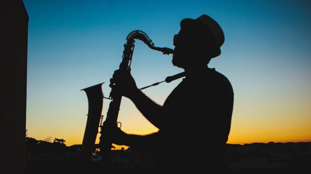 A musician wearing a hat and playing the saxophone with a sunset in the background