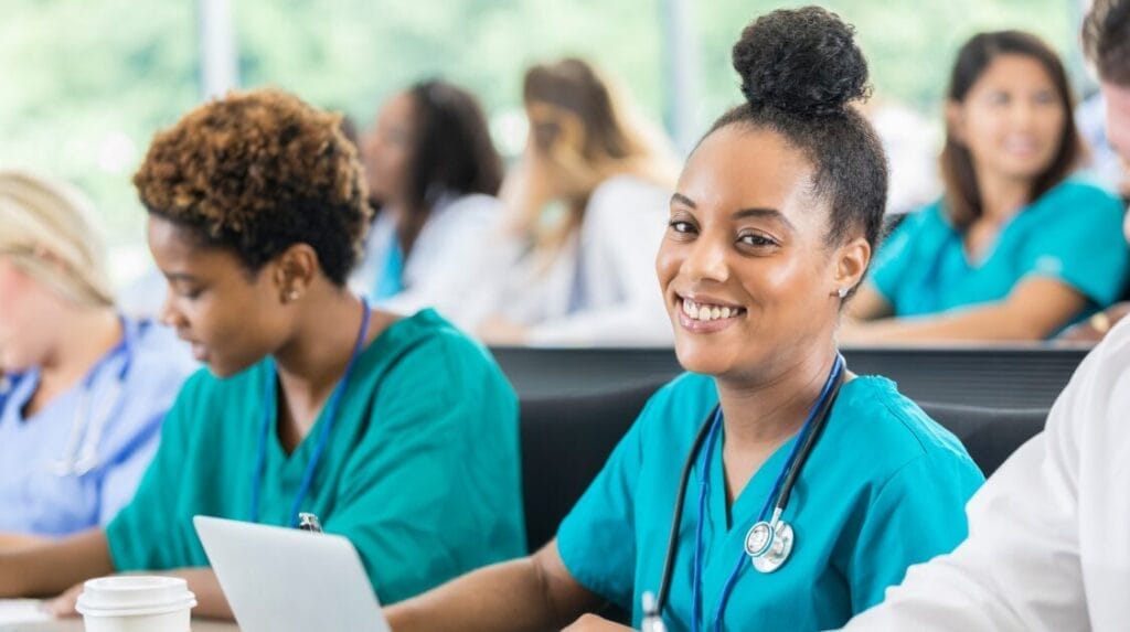 A nurse smiling and sitting at a table with a crowd of nurses in the background