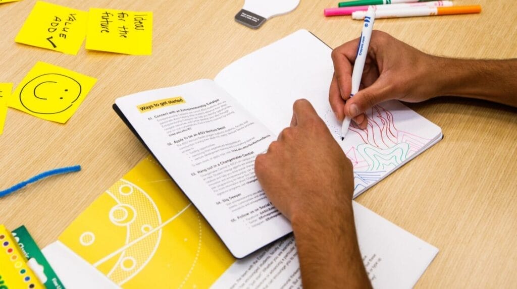 A person writing in the Just Start Guidebook with with sticky notes and markers on the table