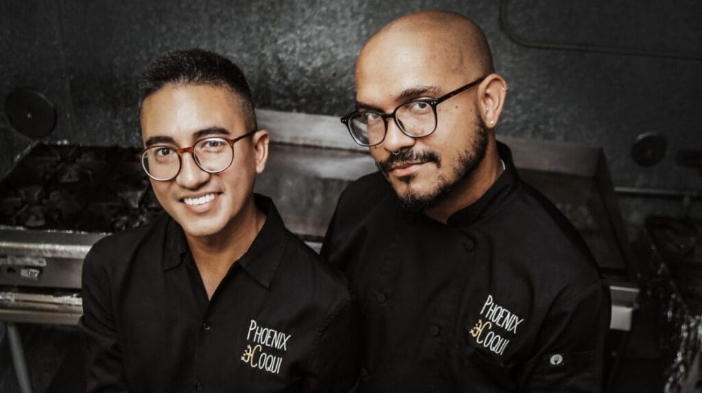 Two people in a kitchen in black chef jackets with Phoenix Coqui embroidered on the jackets
