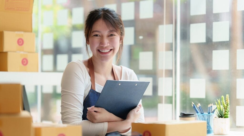 A business owner smiling towards the camera holding a clipboard with shipping boxes stacked in the background