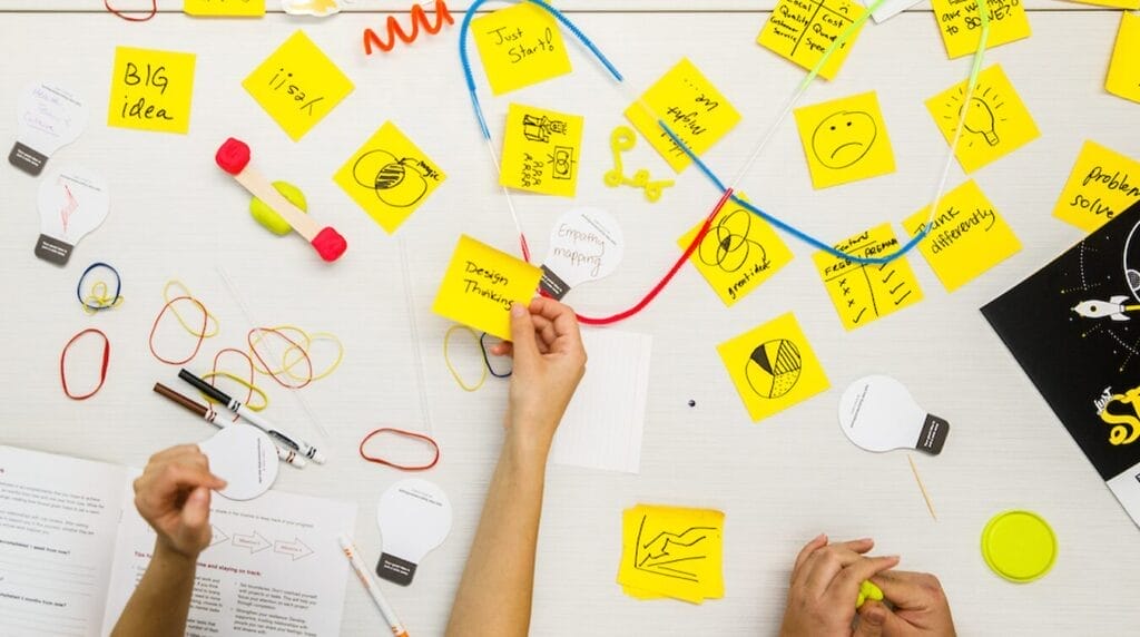A table of bright colored sticky notes and hands reaching for the notes