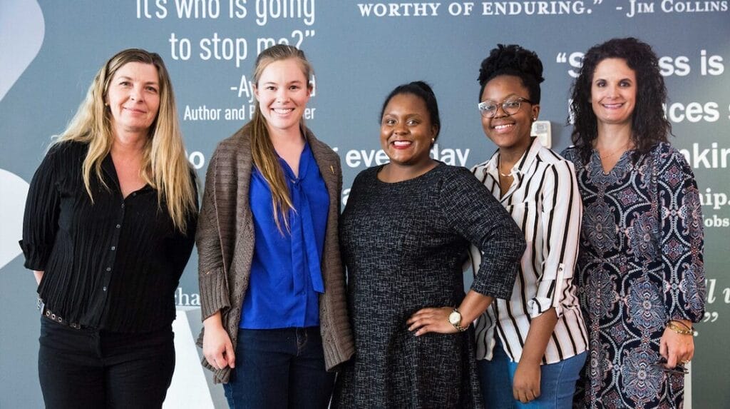 Five professional people standing in front of a wall with text on it and smiling at the camera