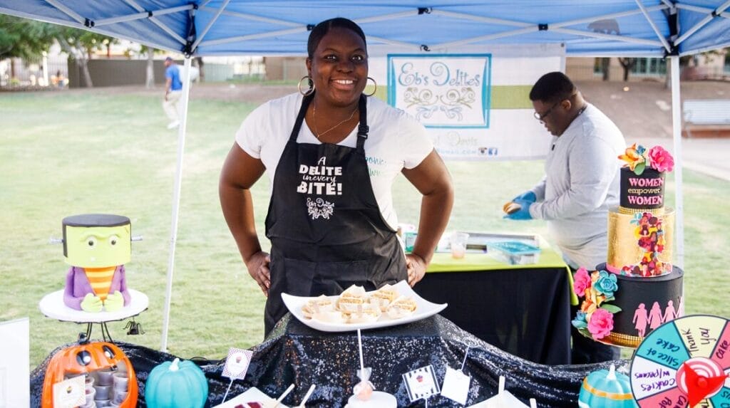 A person smiling at the camera wearing an apron and selling her gourmet desserts at an outdoor market