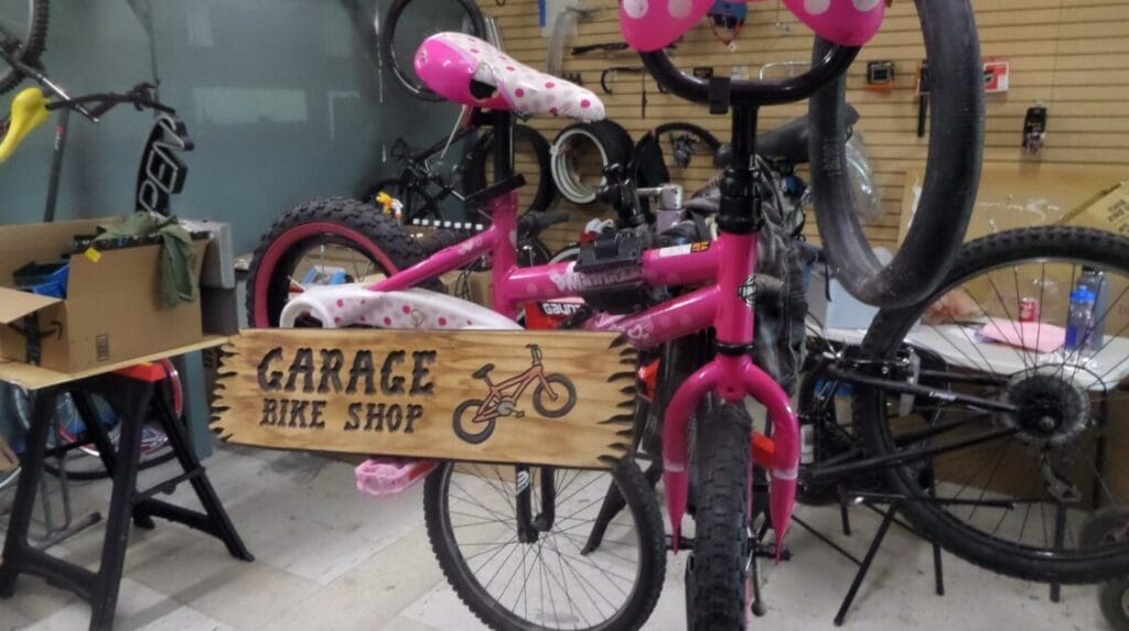 A pink bike with a wood sign hanging from it that reads “Garage Bike Shop”