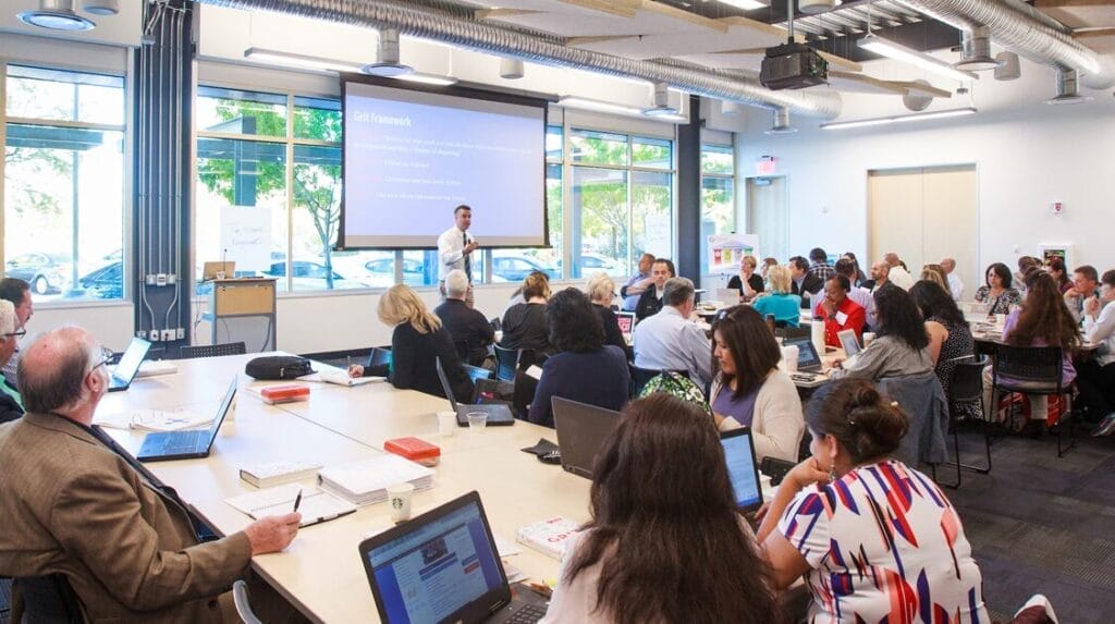 A group of people sitting at tables watching a person speaking in front of a screen with a window and trees behind the screen