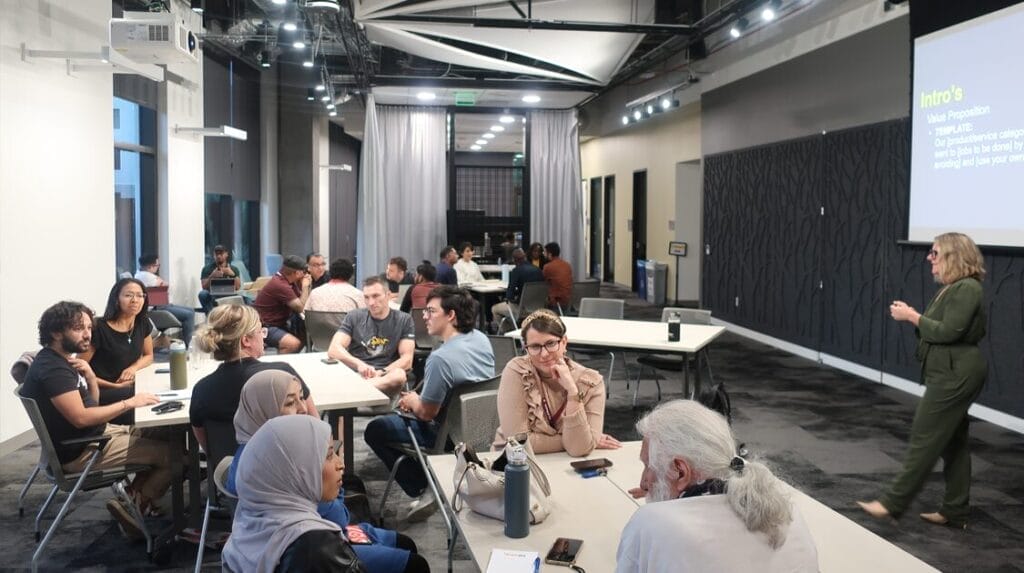 A group of people sitting at tables watching a person speaking in front of a screen