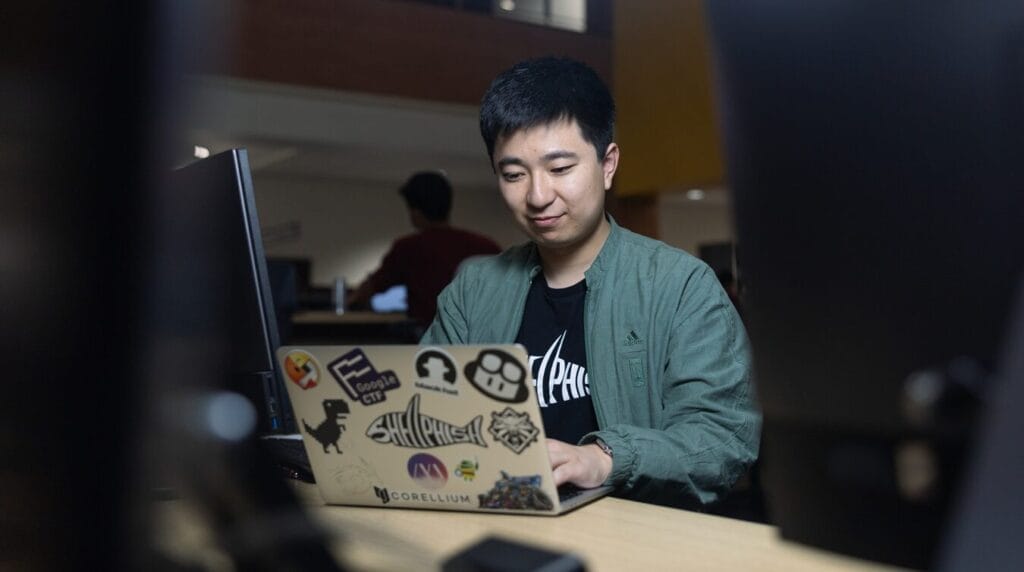 Person sitting in a dark community room typing on a laptop with stickers all over the laptop exterior