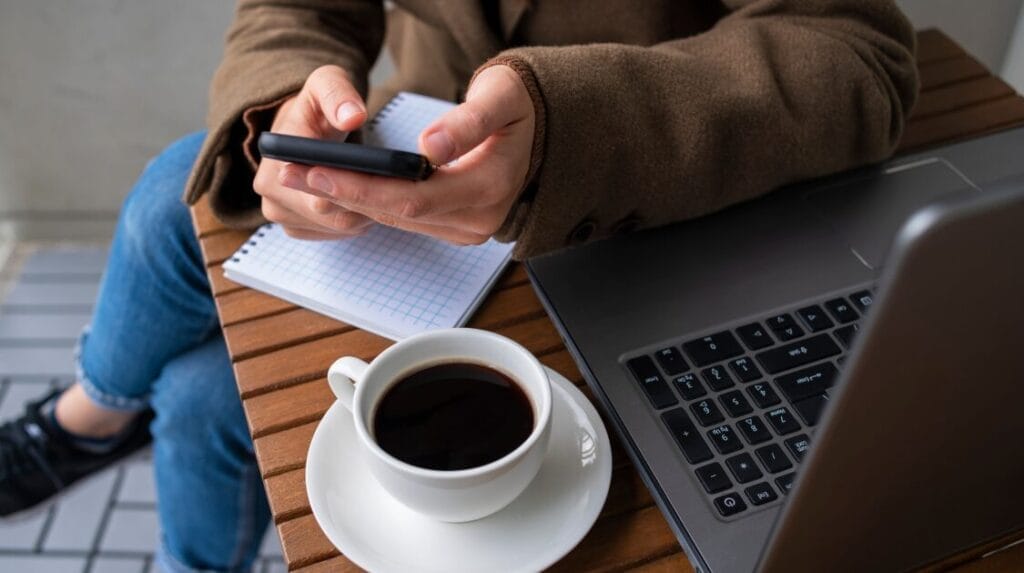 Hands holding a mobile phone next to a coffee cup, a laptop and a notebook