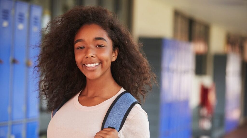 High school student holding her backpack strap, smiling at the camera and standing in front of school lockers