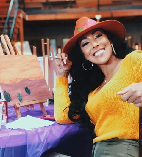 Acacia Barnett smiling and wearing a hat sitting at a table with a painting on a small easel