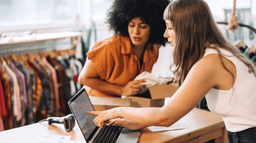 Two store owners looking at a laptop screen with a rack of clothing in the background