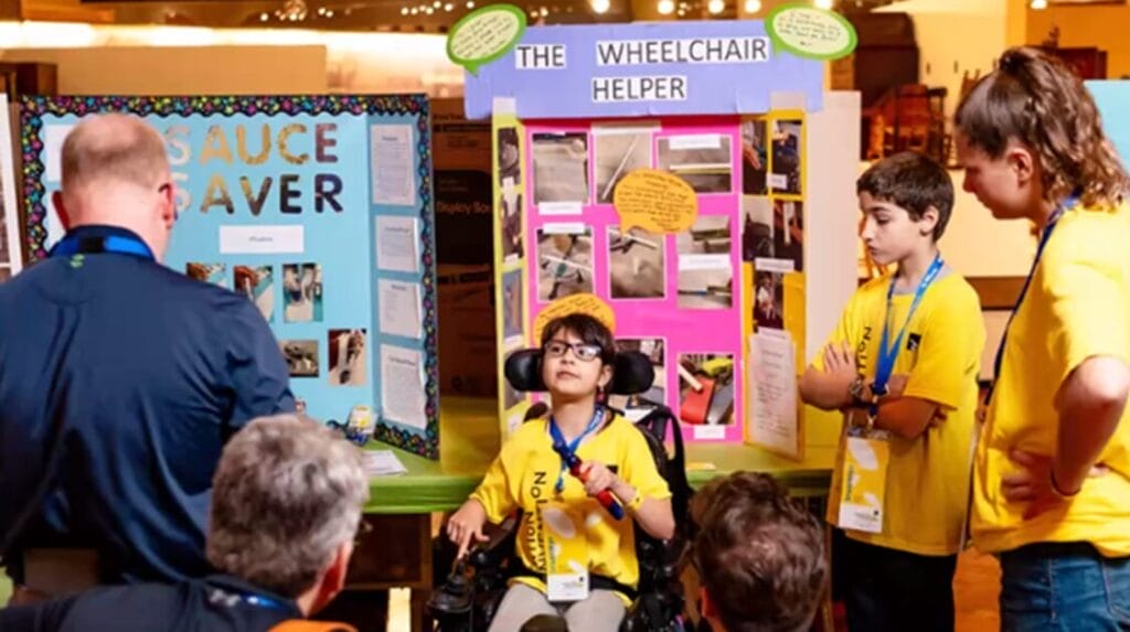 A child sitting in front of their presentation talking to a small audience about their invention called “The Wheelchair Helper.”