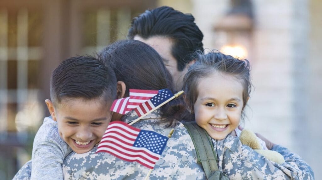 A person in the military hugging their three family members and holding two small American flags