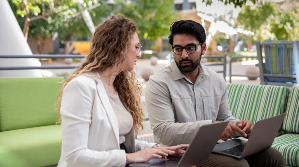 Two professional people sitting outdoors with laptops on their laps and talking to each other
