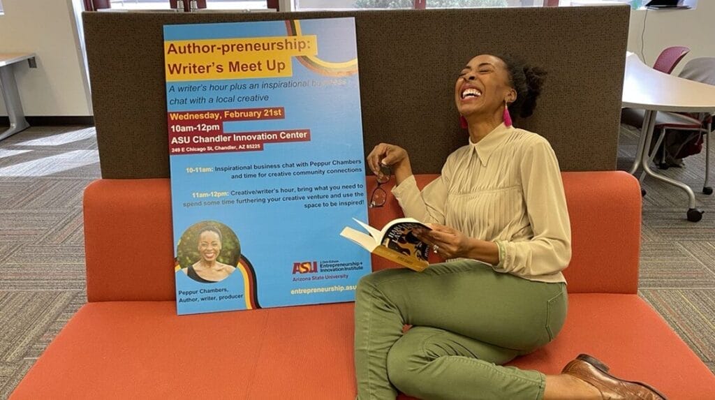 A person sitting next to a sign for an Author-preneurship event and laughing while holding a book.
