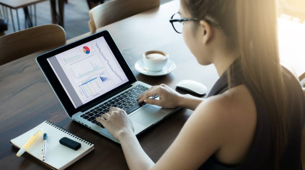 A person wearing glasses, sitting at a table, typing on a laptop with graphs on the screen