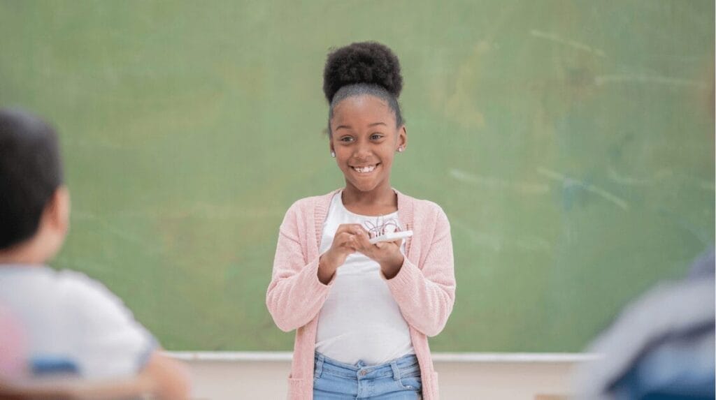 A child smiling and presenting her project to her classroom