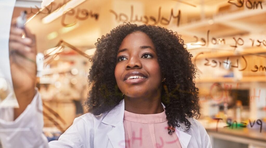 A college student wearing a lab coat, smiling and looking at a window she is writing on with a marker