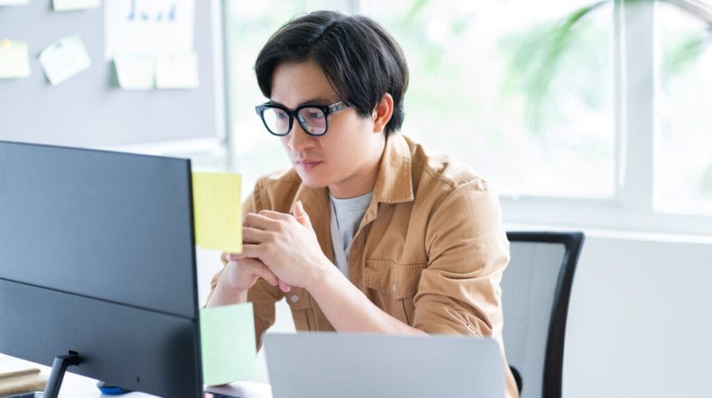 A college student wearing glasses sitting in front of a computer screen with a window in the background